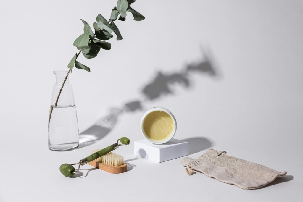 Aesthetic studio shot of skincare products including a jade roller, brush, and cream jar with eucalyptus branch.